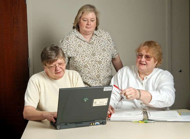 Three people are engaged in a collaborative setting, gathered around a laptop. Two are seated at a table, looking at the screen, while another stands nearby, observing. Papers and highlighters are spread on the table, indicating a work or learning environment.