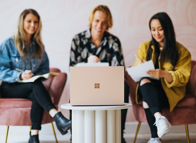 Three people are seated on pink chairs around a small, modern circular table with a laptop placed on top. They appear to be engaged, taking notes and smiling, suggesting a collaborative or meeting environment.