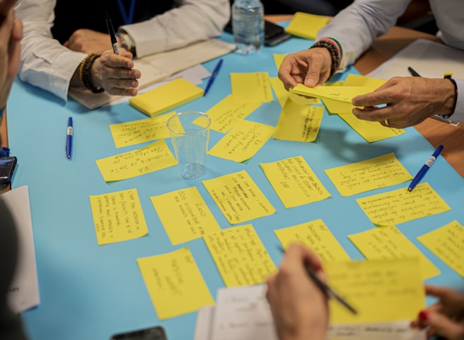 A group of people participate in a collaborative brainstorming session. Yellow sticky notes are scattered across a blue table, each containing handwritten ideas or notes. Several hands are in motion, holding pens and sticky notes, indicating active participation. A bottle of water and a clear plastic cup are also on the table.