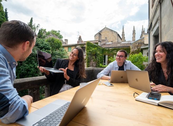 A group of four people sit around a wooden table, each with a laptop and one person holding a tablet. They are engaged in a lively discussion, smiling and appearing collaborative. The setting is outdoors on a patio with greenery and historic buildings in the background.