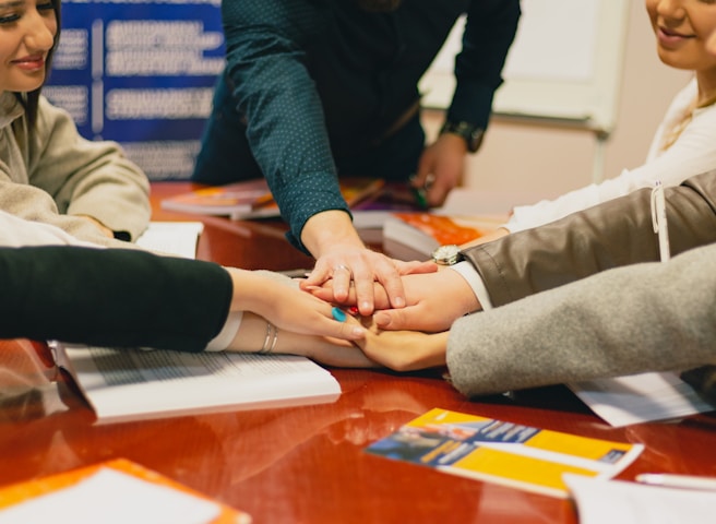 Several people gather around a table placing their hands on top of each other, symbolizing teamwork or unity. The scene is set in an indoor environment with a focus on collaboration. Documents and open books are visible on the table, suggesting a meeting or group study session.