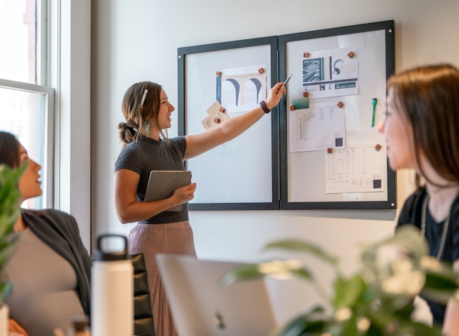 A group of people are engaged in a collaborative meeting in an office setting. A woman stands pointing at a whiteboard with drawings and notes while holding a tablet. Two other women are seated, facing the standing woman, and appear to be attentively listening. The room is well-lit through a large window, and there are some plants and office supplies visible on the table.