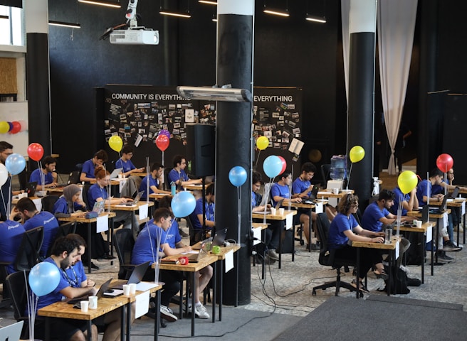 A group of people dressed in blue shirts sit at tables in a modern, open-space office environment. They are focused on their laptops, with paperwork and personal items scattered on desks. Balloons of various colors are tied to the tables, and a large projector hangs from the ceiling. The backdrop displays a wall with the words 'Community is Everything' and photo collages.